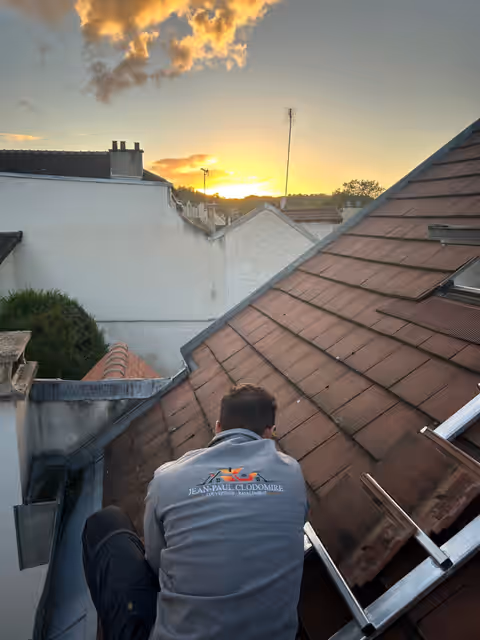 Jean-Paul Colombus de dos sur une toiture en tuiles, veste brodée JPC Couverture, au coucher du soleil
